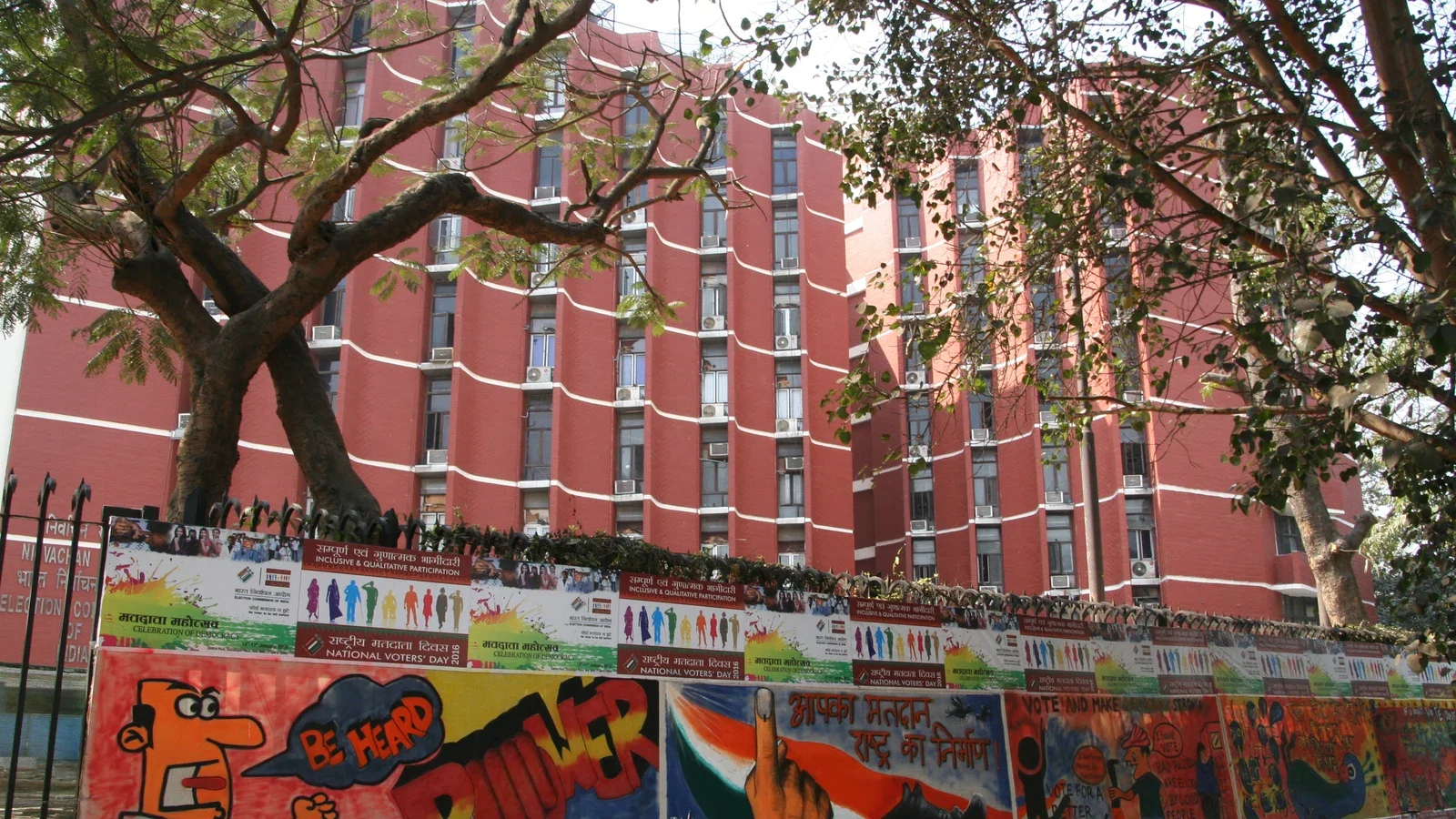 Red-brick Election Commission of India office blocks in New Delhi seen through tree branches, behind a wall of National Voters' Day posters from 2016.