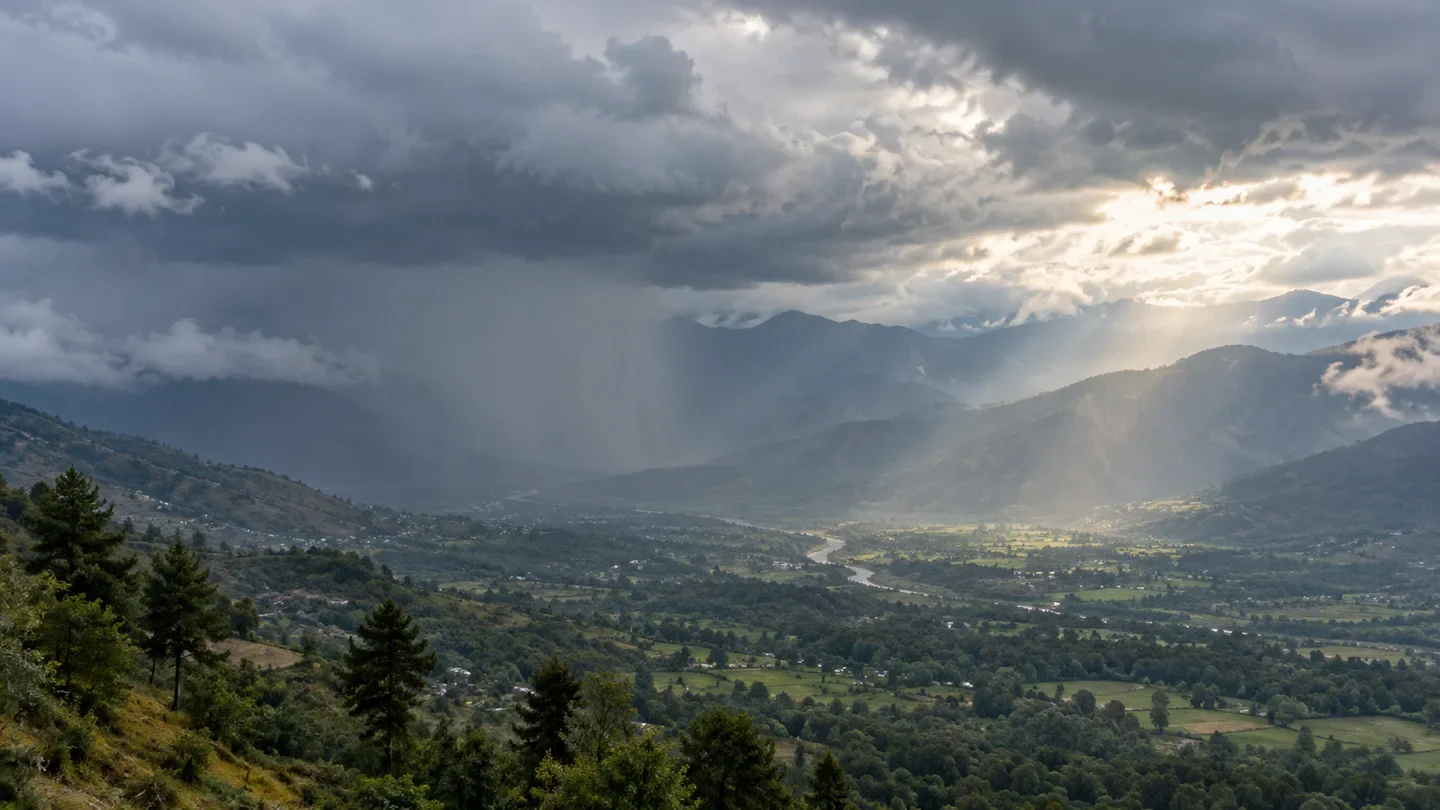 Layered storm clouds and a band of rain over a green valley and distant mountain ridges, with sunlight breaking through nearby.