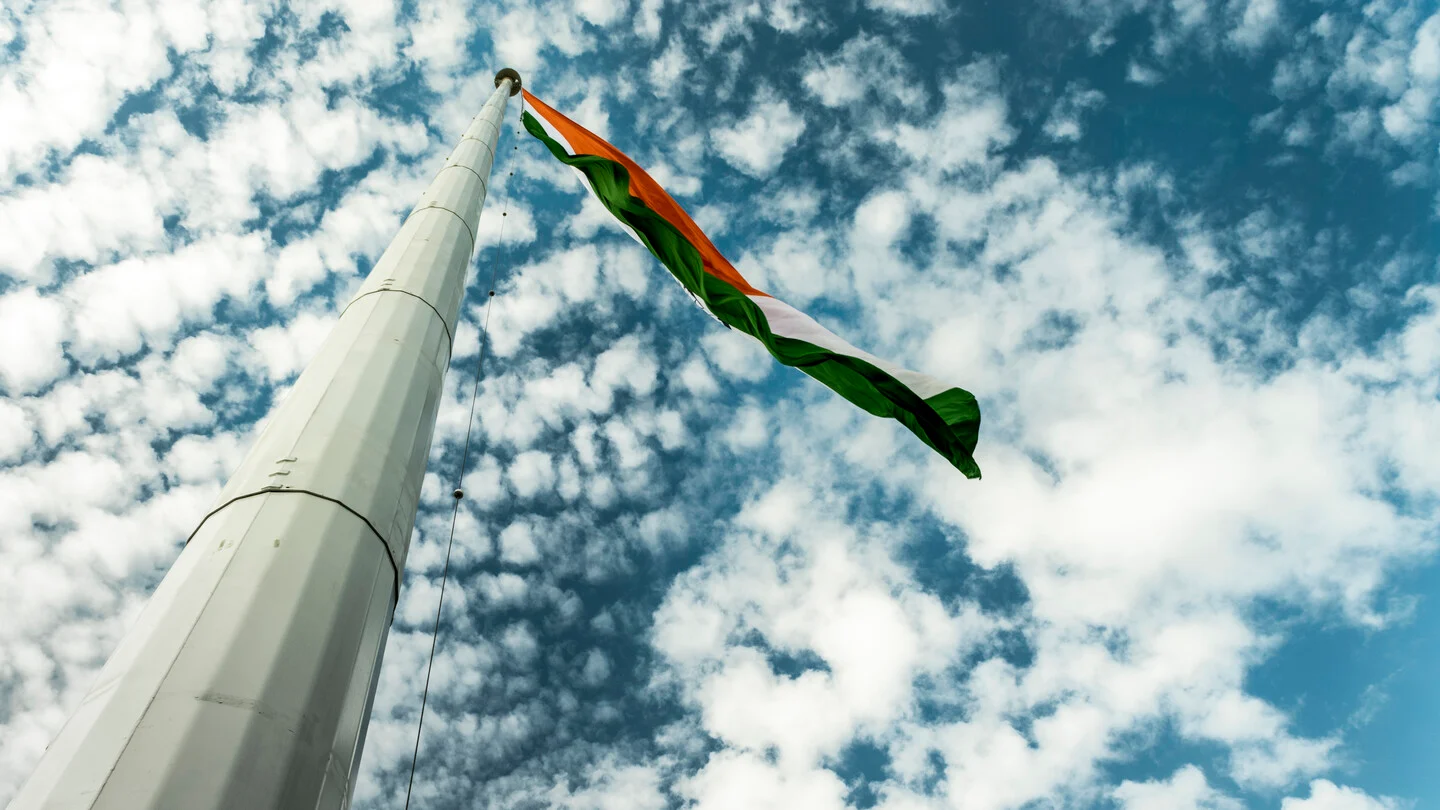 A tall white flagpole seen from below, flying the saffron, white and green Indian flag against a blue sky with scattered white clouds.