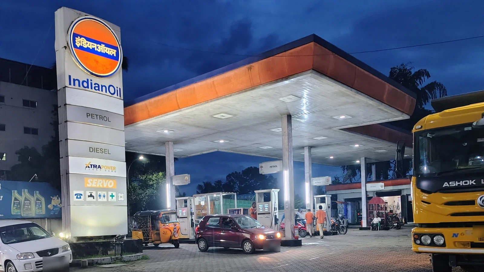 A lit Indian Oil fuel station at dusk with a tall sign listing petrol and diesel, a canopy over the pumps, a few cars and a yellow truck.