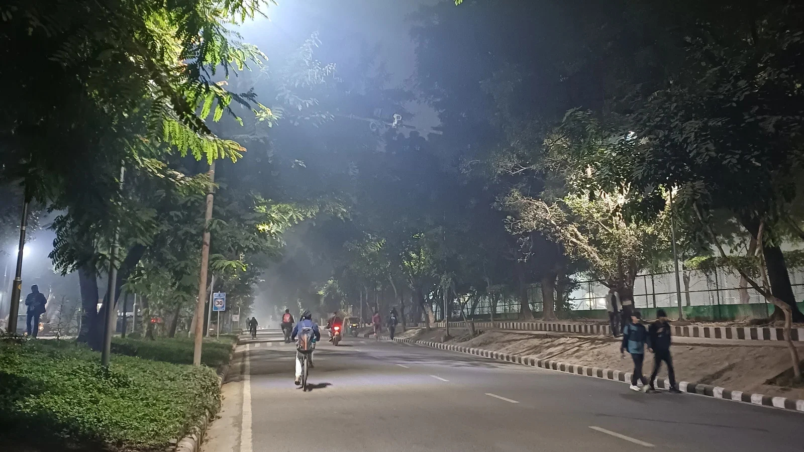 A tree-lined road at night under hazy air, with a streetlight glowing through the haze above a cyclist, motorbikes and pedestrians.