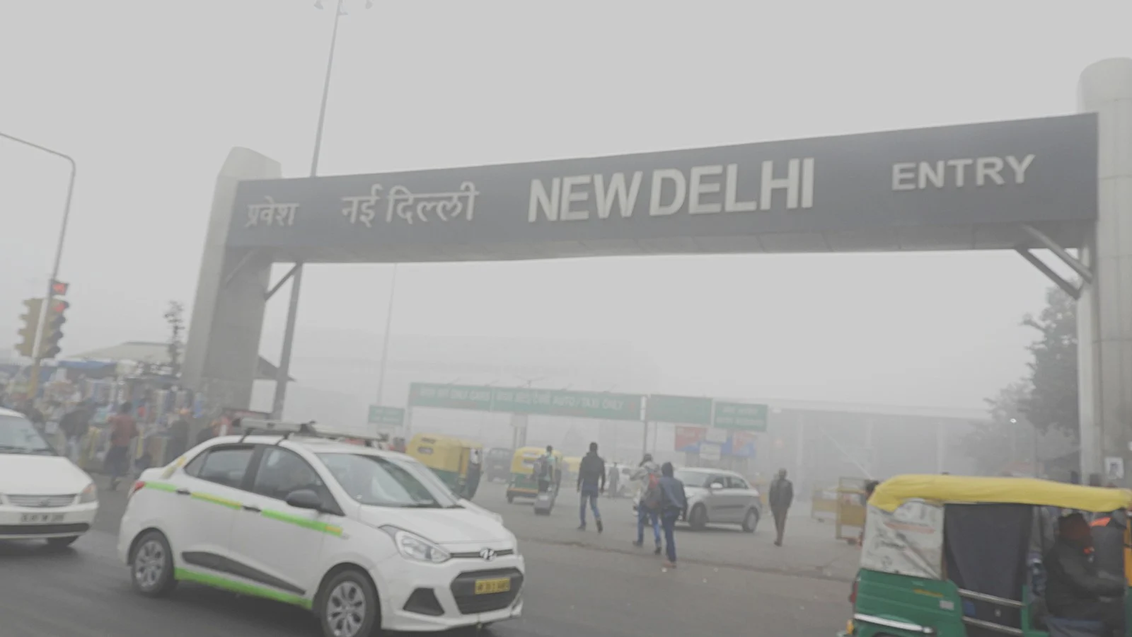 A white taxi and a green-and-yellow auto-rickshaw pass in front of the "New Delhi" entry gate at New Delhi railway station, with pedestrians and distant vehicles faint in thick grey smog.